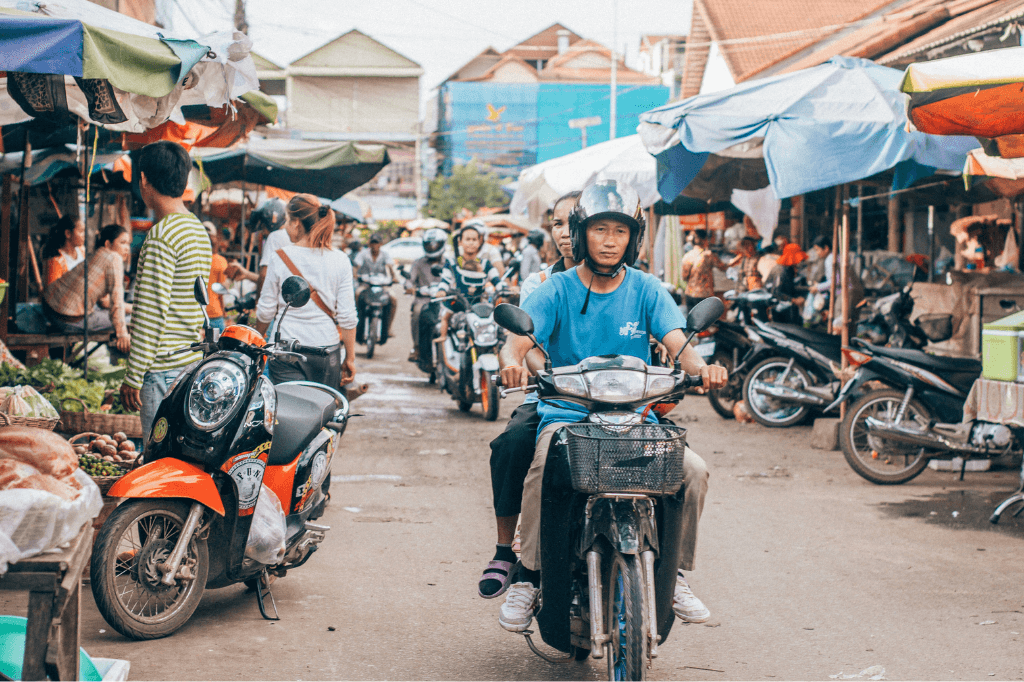 Man riding a motorcycle with a woman on the back, passing through a busy Asian market